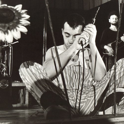 André Herzberg, bekleidet mit einer gestreiften Latzhose und einer Clownsnase im Gesicht, sitz auf dem Bühnenboden. ER hat den Kopf gesenkt und die Aiugen geschlossen. In den Händen hält er eine Bohrmaschine. Mit dieser hat e Löcher in ein Brett gebohrt und dann künstliche Sonnenblumen hineingesteckt. Im Hintergrund steht der Bassist von PANKOW mit seinem Instrument.